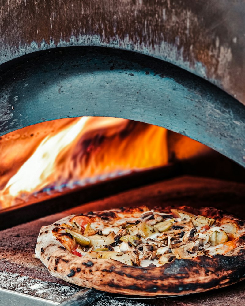 Pizza vor der Öffnung eines Holzofens, dahinter lodert das Feuer