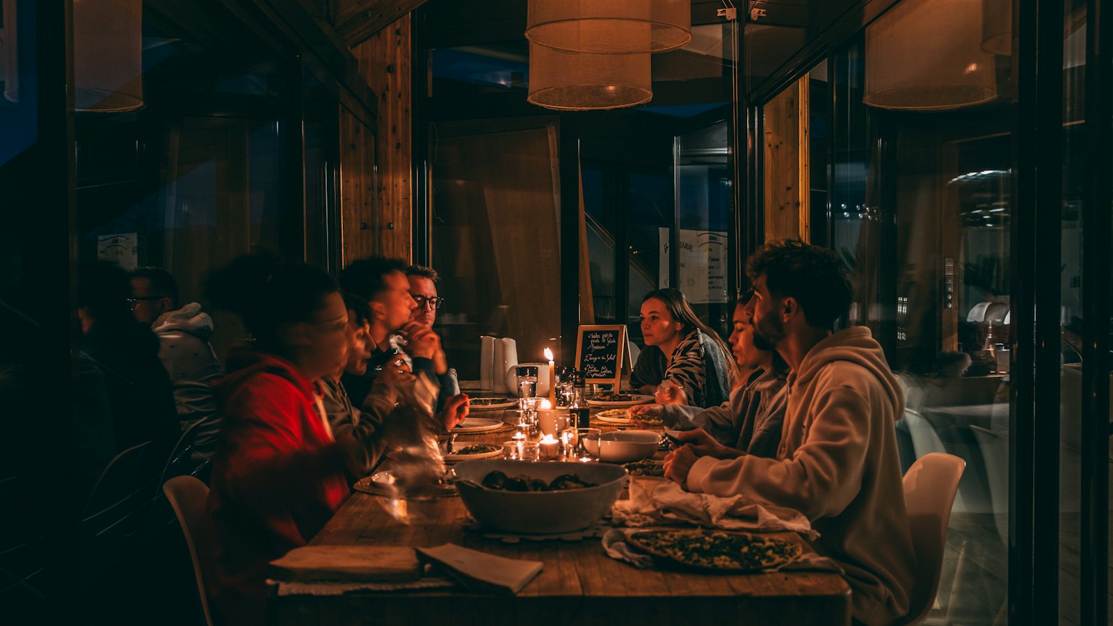 Gäste an einer langen Tafel bei Kerzenlicht in einem dunklen Gastraum