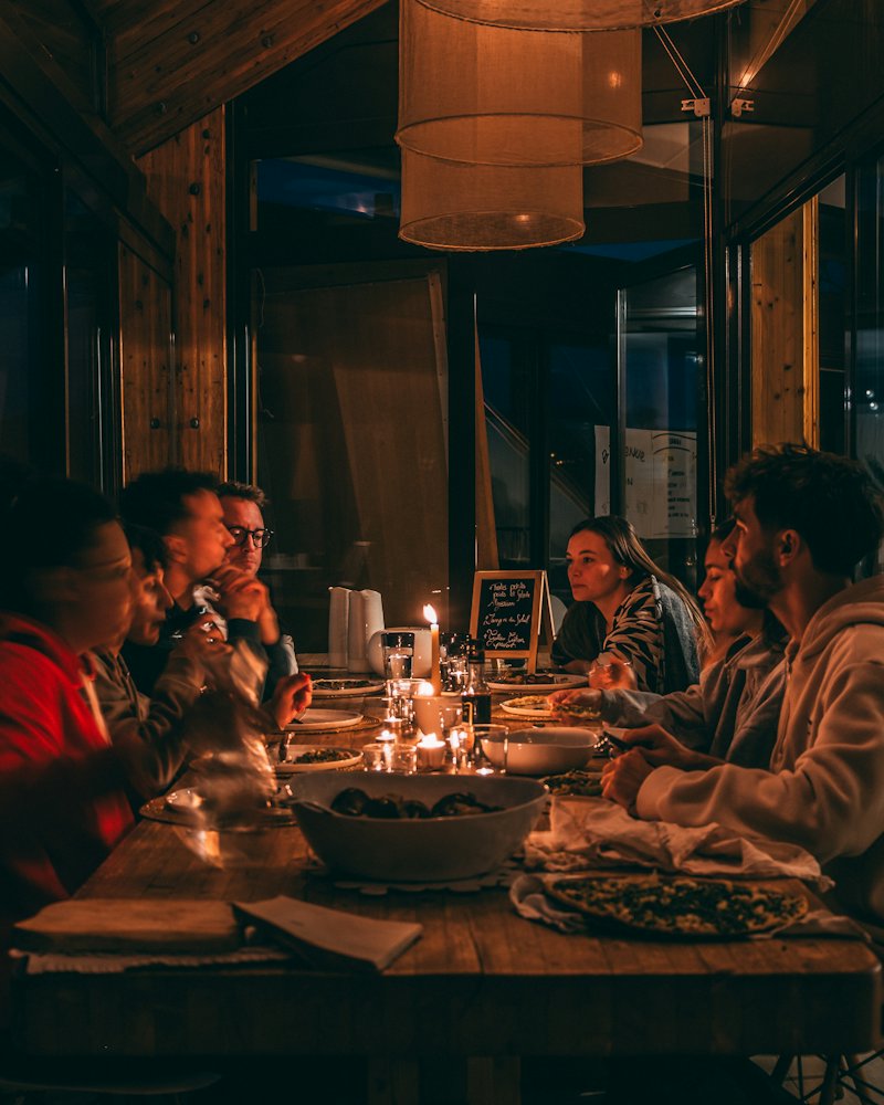 Gäste an einer langen Tafel bei Kerzenlicht in einem dunklen Gastraum